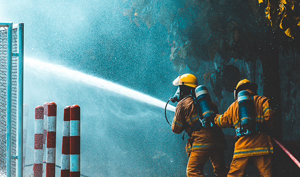 Two firefighters in protective gear spraying water from a fire hose, with water misting into the air, and red and white striped poles in the foreground.