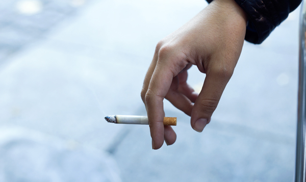 A hand holding a lit cigarette with smoke rising from the tip, set against a blurred background.