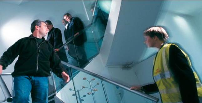 A group of people walking down a staircase, with one person wearing a yellow reflective vest, possibly acting as a fire warden, and others in regular clothing.