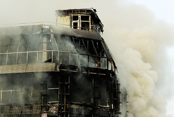 A building heavily damaged by fire, with smoke rising from the structure and windows blackened from the blaze.