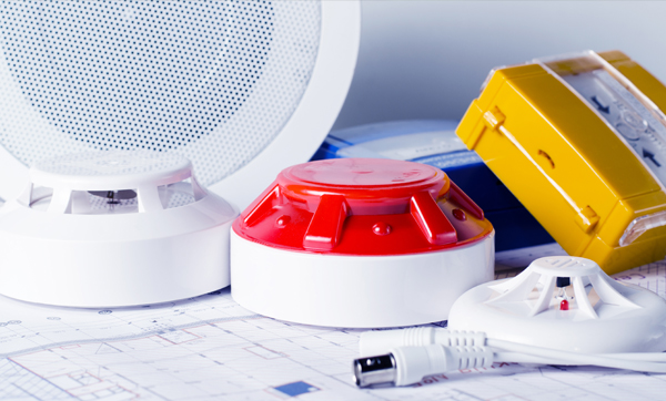 A collection of fire safety devices including smoke detectors, a red alarm bell, and a yellow alarm box, placed on top of blueprints.