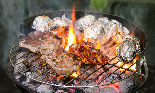A close-up of meat sizzling on a barbecue grill alongside foil-wrapped potatoes, with flames and smoke rising from the hot coals.