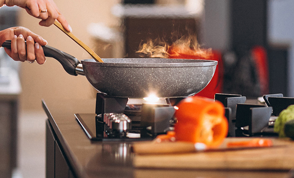 A person stirring food in a frying pan on a stove with visible flames and smoke, surrounded by fresh vegetables on a kitchen countertop.
