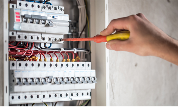 A hand holding a screwdriver while working on an electrical circuit board, with various wires and breakers visible in the electrical panel.