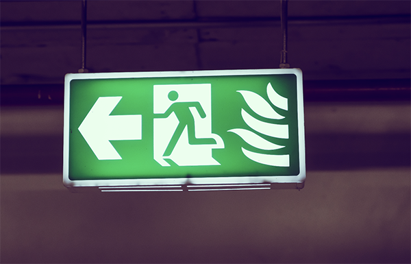 An emergency exit sign showing a person running towards an exit with a left arrow and flames, indicating a fire escape route.