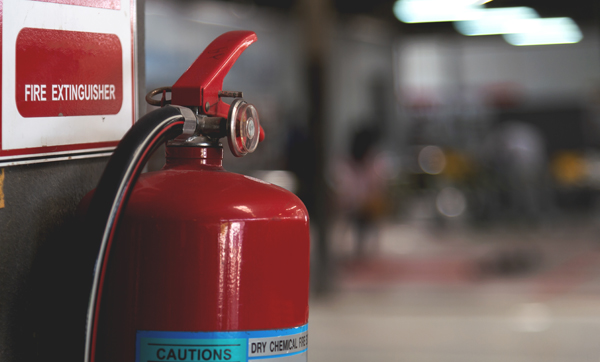 A close-up of a red fire extinguisher mounted on the wall with a caution label, and a blurred background of an industrial setting.