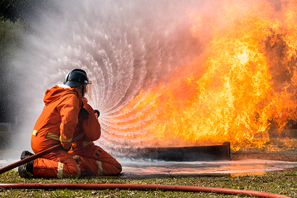 A firefighter in protective gear spraying water on a large fire, using a fire hose, with water creating a spiral pattern as it hits the flames.