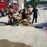 Firefighters in full gear push a person on a stretcher across a concrete surface near a fire truck and brick building. A person in a black uniform assists. The scene appears to be a training or emergency response setting.