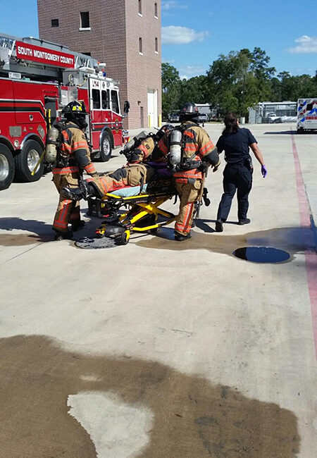 Firefighters in full gear push a person on a stretcher across a concrete surface near a fire truck and brick building. A person in a black uniform assists. The scene appears to be a training or emergency response setting.