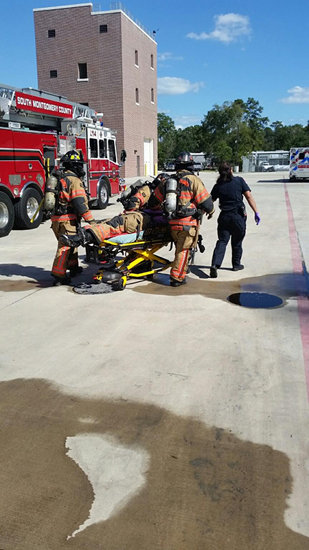 Firefighters in full gear push a person on a stretcher across a concrete surface near a fire truck and brick building. A person in a black uniform assists. The scene appears to be a training or emergency response setting.