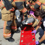 Two firefighters in gear help a woman, held upside down, off a red backboard on a concrete surface. A paramedic assists at the end of the board, wearing a blue uniform and purple gloves. A red emergency vehicle is visible in the background.