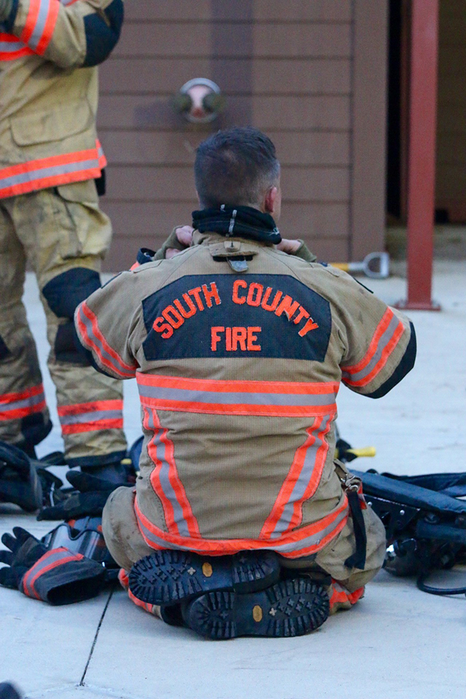 A firefighter sits on the ground, facing away, wearing gear labeled South County Fire. He is surrounded by equipment. Another person in firefighting gear is partially visible to the left.
