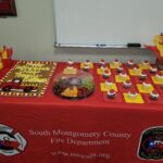 A decorated table features a sheet cake with a toy fire truck, cupcakes, a fruit tray, and red and yellow decor. The tablecloth displays South Montgomery County Fire Department with logos and the website mcesd8.org.