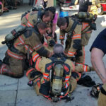Firefighters in gear assist a sitting colleague at a fire station. Several firefighters gather around, ensuring their colleague is okay. Equipment and a fire truck labeled South Montgomery County are visible in the background.