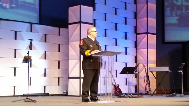 A person in a formal uniform stands at a clear podium on a stage with geometric patterned walls. Musical equipment and a microphone stand are nearby, and large screens display partial images in the background.