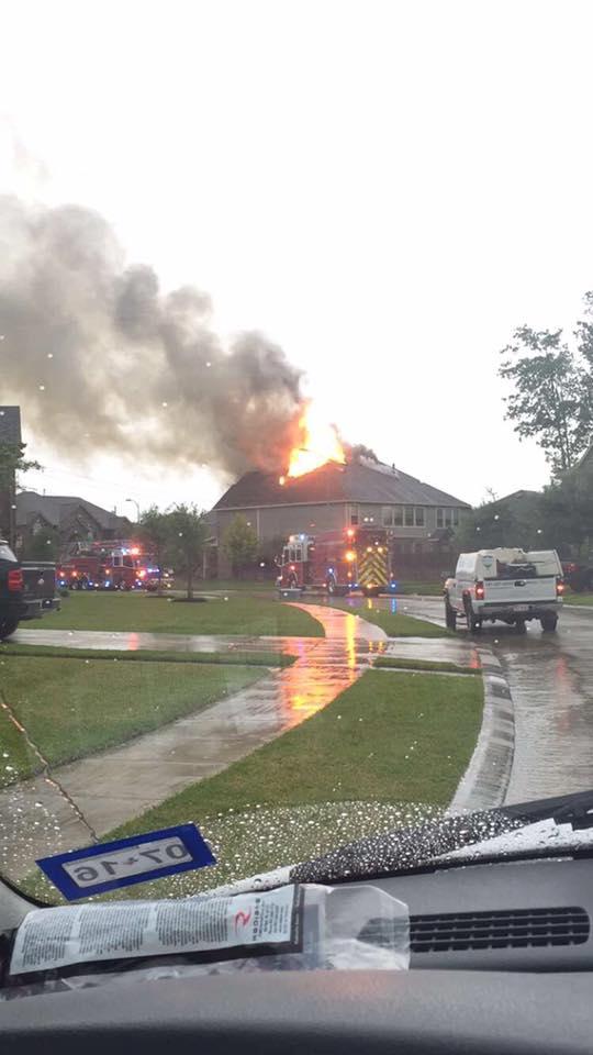 A house is on fire with flames and smoke rising from the roof. Several fire trucks and emergency vehicles are parked on the street. The road is wet, indicating recent rain. A vehicle is in the foreground, viewed from inside another car.