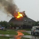 A house with flames and smoke rising from the roof. Fire trucks and emergency vehicles are on the street. The sky is overcast, and the ground is wet from rain. Trees and nearby houses are visible.