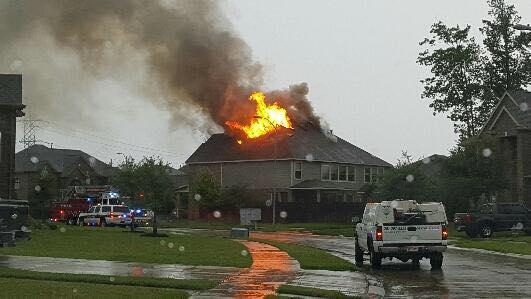 A house with flames and smoke rising from the roof. Fire trucks and emergency vehicles are on the street. The sky is overcast, and the ground is wet from rain. Trees and nearby houses are visible.