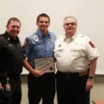 Three uniformed men stand together. The man in the middle holds a Certificate of Appreciation. They wear badges and patches indicating their firefighting roles. Smiles are on their faces, suggesting a celebratory moment.