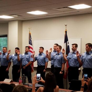 Nine uniformed firefighters stand in a line with hands raised for an oath-taking ceremony. They are facing forward with an American flag and another flag behind them. People in the foreground hold up phones to capture the moment.