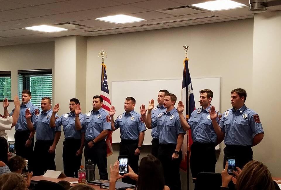 Nine uniformed firefighters stand in a line with hands raised for an oath-taking ceremony. They are facing forward with an American flag and another flag behind them. People in the foreground hold up phones to capture the moment.