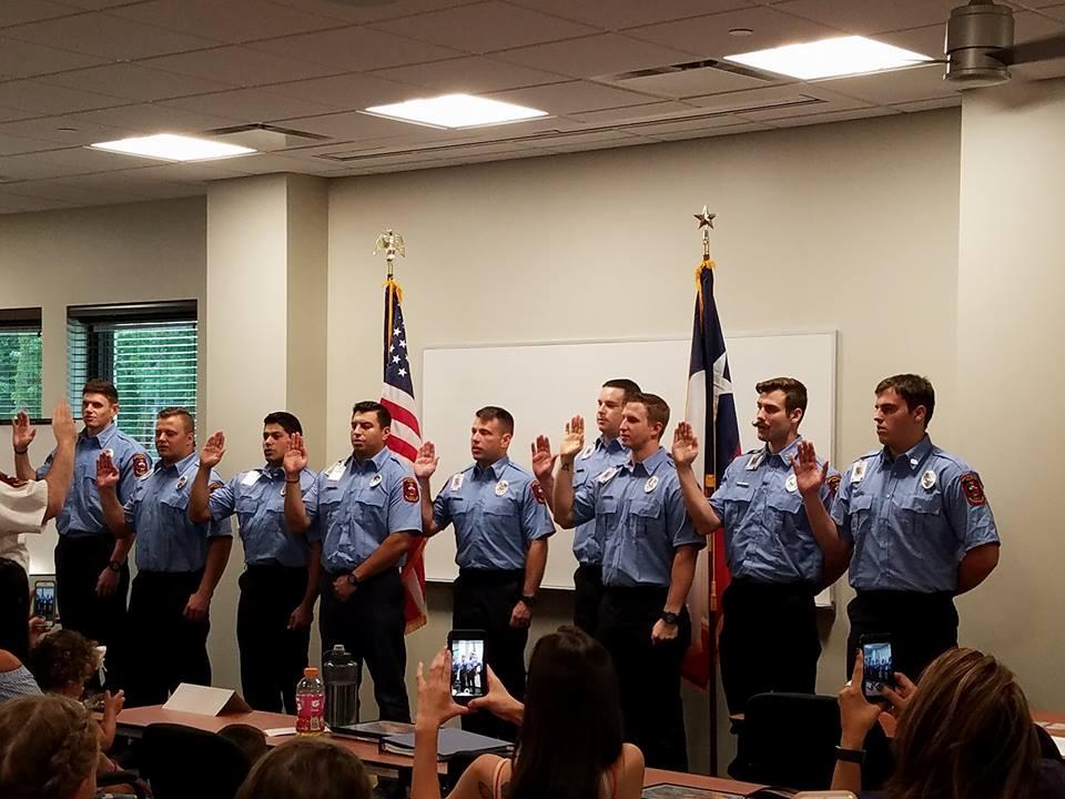 Nine uniformed firefighters stand in a line with hands raised for an oath-taking ceremony. They are facing forward with an American flag and another flag behind them. People in the foreground hold up phones to capture the moment.