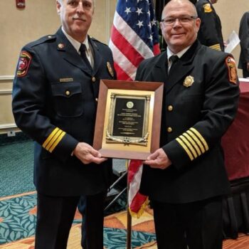 Two uniformed men stand indoors, holding a plaque together. An American flag is behind them. Both are wearing dark formal attire with badges. A few people are in the background. The setting appears to be an award ceremony.