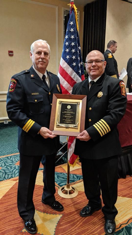 Two uniformed men stand indoors, holding a plaque together. An American flag is behind them. Both are wearing dark formal attire with badges. A few people are in the background. The setting appears to be an award ceremony.