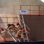 Firefighters in full gear climb ladders at a training facility. They are surrounded by smoke, working together near a structure labeled Dräger Training System. The scene focuses on emergency response practice.