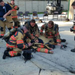 Firefighters in gear are gathered on a concrete floor, attending to a colleague who is lying down. Some chairs and a forklift are in the background. Another firefighter stands nearby, observing the scene.