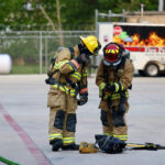 Two firefighters in full gear are preparing equipment in an outdoor setting. One is adjusting a tool while the other looks on. They are near a chain-link fence and a fire safety trailer. A hose is visible on the ground.