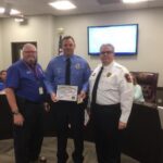 Three men in a conference room. Two are wearing blue shirts; one in the center is holding a certificate. The man on the right is in a white uniform. A large screen and flags are in the background.