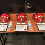 Three firefighter helmets labeled Wilson, Mueller, and Steel are on a table with certificates of promotion and identification cards beneath them. The backdrop features a wooden door and a dark wall.