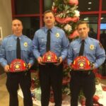 Three firefighters in uniform stand holding red helmets in front of a decorated Christmas tree with red and white ribbons. They are indoors, and Christmas presents can be seen beneath the tree.