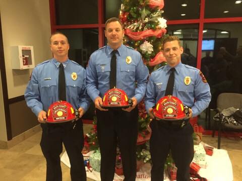 Three firefighters in uniform stand holding red helmets in front of a decorated Christmas tree with red and white ribbons. They are indoors, and Christmas presents can be seen beneath the tree.