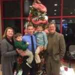 A family of five poses in front of a decorated Christmas tree. Two adults, a man and a woman, stand on either side. A man holds two young boys, one in each arm. All are smiling and dressed in semi-formal attire.