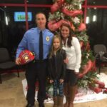 A man in a blue uniform holds a firefighters helmet, standing beside a woman and a young girl in front of a decorated Christmas tree with red ribbons. They are smiling and indoors, possibly at a holiday event.