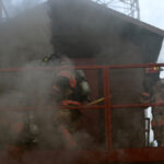 Firefighters in full gear approach a smoke-filled building. Two are on a balcony, one carrying a tool, while thick smoke billows around them. Safety equipment and teamwork are evident in their coordinated efforts.