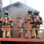 Four firefighters in full gear, equipped with oxygen tanks, stand on an outdoor structure surrounded by smoke. They appear to be entering a doorway in a training exercise or emergency scenario. Trees and a metal tower are visible in the background.