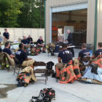 A group of firefighters in uniform sit and stand around a tall garage. Some are wearing firefighting gear. Equipment and tools are visible on the ground. One person appears to be addressing the group, while another takes a photo. Trees are in the background.