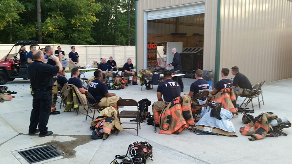 A group of firefighters in uniform sit and stand around a tall garage. Some are wearing firefighting gear. Equipment and tools are visible on the ground. One person appears to be addressing the group, while another takes a photo. Trees are in the background.