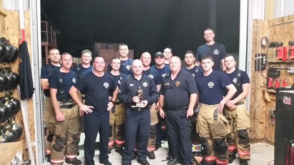 A group of firefighters, wearing uniforms and some with helmets, stand in a garage. One holds a trophy. They appear to be inside a fire station, with various equipment visible around them. The atmosphere is relaxed and camaraderie is evident.