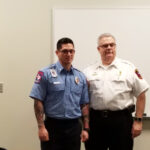 Two men in uniform stand side by side in front of a whiteboard. The man on the left wears a blue uniform with tattoos on his arms, while the man on the right wears a white uniform. Both have badges on their shirts.