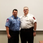 Two uniformed men stand side by side in front of a blank white wall. The man on the left wears a blue uniform, while the man on the right wears a white uniform. They are posing for the photograph in what appears to be an indoor setting.