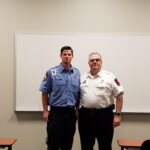 Two men in uniform stand side by side in front of a whiteboard. One wears a blue shirt with badges; the other wears a white shirt with a badge. Both are smiling. Desks and a partial view of an audience are visible in the foreground.