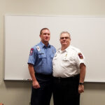 Two men in uniform stand together in front of a whiteboard. One wears a blue shirt with patches, and the other wears a white shirt with insignias. They are smiling and standing in a classroom setting.