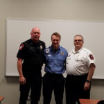 Three men in uniform stand together smiling in a classroom with a whiteboard behind them. The man on the left wears a black uniform, the middle one wears a blue uniform, and the right one is in a white shirt with a badge.