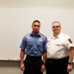 Two men standing indoors in front of a whiteboard. One is wearing a blue uniform shirt, while the other is in a white uniform shirt. Both have badges on their shirts and are looking at the camera, with the man in white smiling.