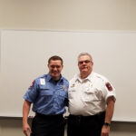 Two men in uniform stand smiling in front of a blank whiteboard. One wears a blue shirt with a badge and the other wears a white shirt with patches. They appear to be in a professional setting.