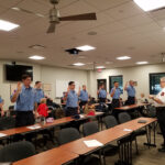 A group of uniformed individuals stands in a classroom with their right hands raised, participating in a ceremony led by an instructor. The room has desks, chairs, and a ceiling with fans, and several onlookers are seated in the background.